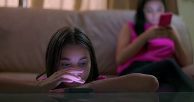 Masaya yaslanan genç bir kız, parlayan akıllı telefon ekranına odaklanmış, çevreden ayrılmış, sosyal izolasyonu ve duygusal kopukluğu gösteren