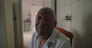 Afrika kökenli yaşlı bir adam fayanslı mutfakta oturuyor, kameraya nazikçe gülümsüyor, sıcaklığını ve bilgeliğini iletiyor, ev ortamında her gün samimi anlar yaşıyor.