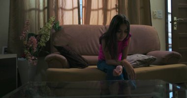 Genç bir kız loş bir odada tek başına oturuyor, kafası eğik, izolasyon, duygusal mücadele ve sakin bir ev ortamında çocukluk bunalımı tasvir ediyor.