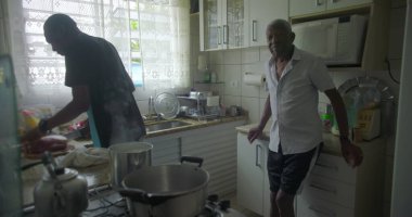 Afrika kökenli bir adam ev mutfağında yemek pişirirken Afrika kökenli yaşlı bir adam yakınlarda durup aile dinamiklerini ve günlük hayattaki kuşak bağlarını inceliyor.
