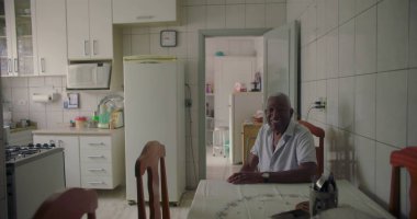 Sıcak bir mutfakta yemek masasında oturan yaşlı Afro-Amerikan bir adam, sıcak bir gülümseme, ev aletleri ile çevrili ve sade bir atmosfer
