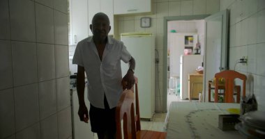 Bir yaşlı siyah adam mutfakta yemek sandalyesinin yanında duruyor, düşünceli görünüyor, mutfak gereçlerinin sade bir geçmişi ve rahat bir iç dekorasyonu var.