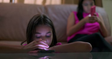 Masaya yaslanan genç bir kız, parlayan akıllı telefon ekranına odaklanmış, çevreden ayrılmış, sosyal izolasyonu ve duygusal kopukluğu gösteren