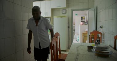 Bir yaşlı siyah adam mutfakta yemek sandalyesinin yanında duruyor, düşünceli görünüyor, mutfak gereçlerinin sade bir geçmişi ve rahat bir iç dekorasyonu var.