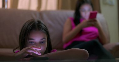 Masaya yaslanan genç bir kız, parlayan akıllı telefon ekranına odaklanmış, çevreden ayrılmış, sosyal izolasyonu ve duygusal kopukluğu gösteren