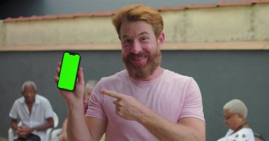 Akıllı telefonu yeşil ekran krom anahtarıyla gösteren adam avluda gülümseyen, arka planda oturan insanlarla dost canlısı ve davetkar bir atmosfer.