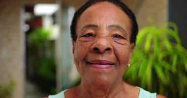 Kameraya sıcacık gülümseyen yaşlı bir Afro-torun kadın. Yüzü, açık havada huzurlu bir ortamda belirtilmiş. Nezaket ve sıcaklık saçıyor..