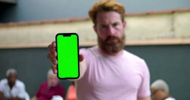 Kameraya doğru yeşil ekran krom anahtarı olan akıllı telefonu uzatan bir adam, günlük avluda, arka planda oturan insanlarla sosyal ortam, düzenleme için yer tutucu ekranı