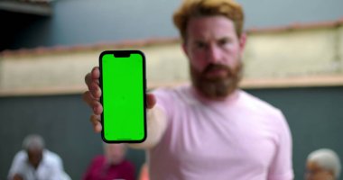 Kameraya doğru yeşil ekran krom anahtarı olan akıllı telefonu uzatan bir adam, günlük avluda, arka planda oturan insanlarla sosyal ortam, düzenleme için yer tutucu ekranı
