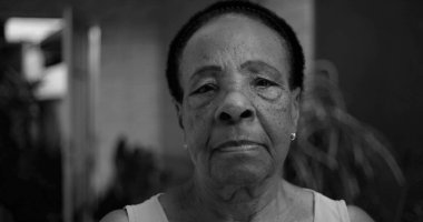 Siyah beyaz bir portre içinde yaşlı bir Afro-soyundan gelen kadın, kameraya ciddi bir ifadeyle bakıyor, yüz hatları açık havada samimi bir evde vurgulanıyor.