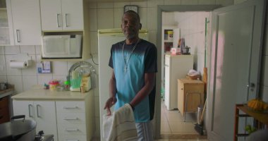 Afrika kökenli bir adam mutfakta elinde havluyla dikiliyor. Düşünceli bir ifade takınıyor. Aile ve aile ortamında sıradan ve samimi bir an.