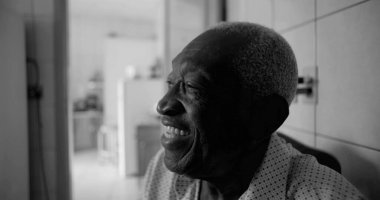 Afrika kökenli yaşlı bir adam fayanslı bir mutfakta neşeyle gülüyor, siyah beyaz bir portre, bir ev ortamında mutluluk, bağlantı ve bilgeliği vurguluyor.