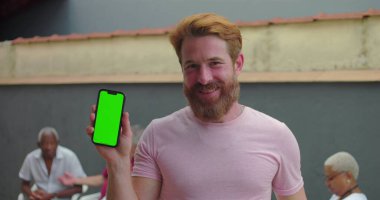 Akıllı telefonu yeşil ekran krom anahtarıyla gösteren adam avluda gülümseyen, arka planda oturan insanlarla dost canlısı ve davetkar bir atmosfer.