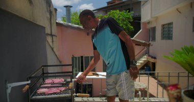 Afrika kökenli Amerikalı bir adam çatıdaki barbeküde et pişiriyor, biftek ve sosisleri kontrol ediyor ve canlı bir şehir ortamında yemek hazırlıyor.