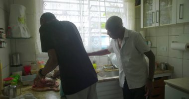 Afrika kökenli yaşlı bir adam elinde neşeli bir ifadeyle bir et parçası tutuyor, kültürel gelenekleri ve aşçılık hazırlıklarını vurguluyor.