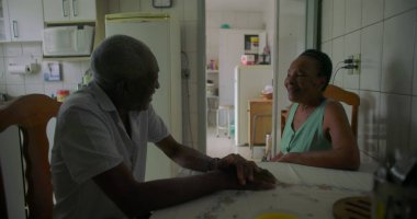 Afrika kökenli yaşlı bir adam ve kadın, bir ev mutfağında bir masada oturuyor, gülümsüyor ve neşeli bir anı paylaşıyor, sıcaklığı ve aile bağlarını vurguluyor.