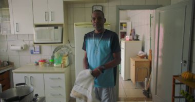 Afrika kökenli bir adam mutfakta elinde havluyla dikiliyor. Düşünceli bir ifade takınıyor. Aile ve aile ortamında sıradan ve samimi bir an.