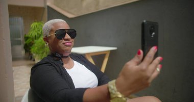 Kendine güvenen bir siyahi kadın görüntülü konuşma yapıyor ya da açık havada bir tanıklığı kaydediyor. Parlak bir şekilde gülümsüyor, arka planda yeşillik olan bir ortamda neşe ve cazibe yayıyor.