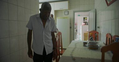 Yaşlı bir siyah adam mutfaktan oturma odasına yürüyor, tarafsız bir ifade takınıyor, ev içinde görünür mobilyalar ve dekorla geçiş yapıyor.