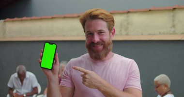 Akıllı telefonu yeşil ekran krom anahtarıyla gösteren adam avluda gülümseyen, arka planda oturan insanlarla dost canlısı ve davetkar bir atmosfer.