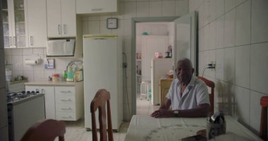 Sıcak bir mutfakta yemek masasında oturan yaşlı Afro-Amerikan bir adam, sıcak bir gülümseme, ev aletleri ile çevrili ve sade bir atmosfer
