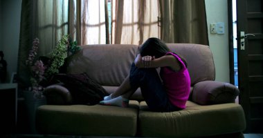 Genç bir kız kanepeye kıvrılmış, kafası kollara gömülmüş, izolasyon, duygusal hassasiyet, çocukluk üzüntüsü ve sakin bir ev ortamında akıl hastalığı gösteriyor.