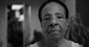 Siyah beyaz bir portre içinde yaşlı bir Afro-soyundan gelen kadın, kameraya ciddi bir ifadeyle bakıyor, yüz hatları açık havada samimi bir evde vurgulanıyor.