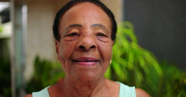 Kameraya sıcacık gülümseyen yaşlı bir Afro-torun kadın. Yüzü, açık havada huzurlu bir ortamda belirtilmiş. Nezaket ve sıcaklık saçıyor..