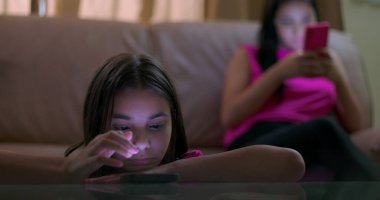 Masaya yaslanan genç bir kız, parlayan akıllı telefon ekranına odaklanmış, çevreden ayrılmış, sosyal izolasyonu ve duygusal kopukluğu gösteren