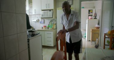 Bir yaşlı siyah adam mutfakta yemek sandalyesinin yanında duruyor, düşünceli görünüyor, mutfak gereçlerinin sade bir geçmişi ve rahat bir iç dekorasyonu var.