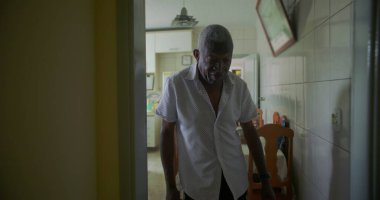 Yaşlı bir siyah adam mutfaktan oturma odasına yürüyor, tarafsız bir ifade takınıyor, ev içinde görünür mobilyalar ve dekorla geçiş yapıyor.