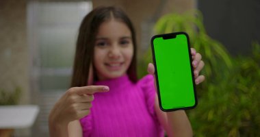 Elinde yeşil ekran krom anahtarı olan akıllı telefonu tutan, neşeli bir ifadeyle boş ekrana bakan, açık bir evde kişiselleştirilebilir içeriği gösteren genç bir kız.