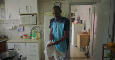 Afrika kökenli bir adam mutfakta elinde havluyla dikiliyor. Düşünceli bir ifade takınıyor. Aile ve aile ortamında sıradan ve samimi bir an.