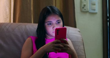 Kanepede oturan genç, yoğun bir şekilde parlak ekranlı akıllı telefona odaklanmış dijital alışkanlıkları, teknoloji kullanımını ve modern iletişim bağımlılığını gösteriyor.