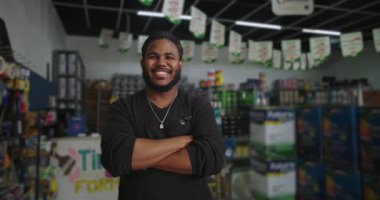 Siyah önlüklü bir adam güvenle gülümsüyor boyacı dükkanında kolları çaprazlanmış, renkli boya malzemeleriyle çevrili, neşeli ve cana yakın davranışlar sergiliyor.