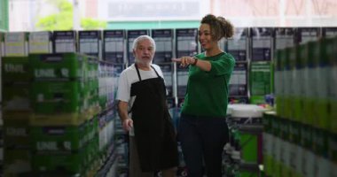 Kıvırcık saçlı yaşlı bir erkek esnaf dükkanında ürün seçeneklerini tartışan, stoklanmış raflardaki eşyaları işaret eden bir kadın müşteri.