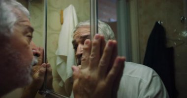 Yaşlı adam yansımasına bakıyor, ağzı şoktan ya da inançsızlıktan hafifçe açık, elleri, dramatik ve duygusal bir kendini anlama anında aynaya bastırılmış.