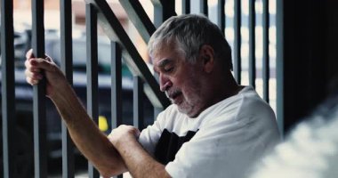 Pencere parmaklıklarını tutan yaşlı bir adam, kafası sessiz bir umutsuzlukla eğik, derin bir duygusal ıstırap ve yalnızlık içinde.