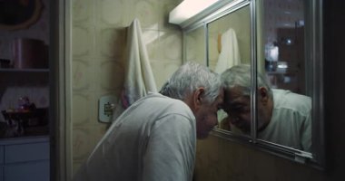 Yaşlı bir adam aynada yüzünü yakından inceliyor, el pençe, derin düşüncelere dalmış ve derin düşüncelere dalmış, eski bir banyoda özel ve yansıtıcı bir an.