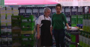 Yaşlı bir erkek esnaf, hırdavatçı koridorundaki kıvırcık saçlı ve içi eşya dolu raflarla çevrili gülümseyen bir kadın müşteriyle etkileşime geçiyor.