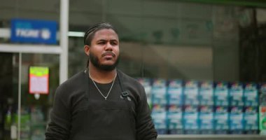 Perakende mağazasının önünde duran odaklanmış genç bir siyahi adam, düşünceli bir şekilde uzaklara bakıyor, iş ortamında profesyonelliği, kararlılığı ve hırsı iletiyor.