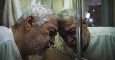 Yaşlı adam alnını aynaya yaslıyor, gözleri kapalı, tükenmişliği, umutsuzluğu ve yalnızlığı yansıtan duygusal ve düşünceli bir anda savunmasızlığı anlatıyor.