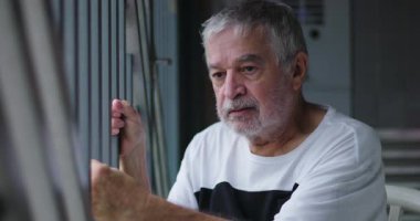 Yaşlı adam oturmuş parmaklıkları tutuyor, ciddi bir ifadeyle dışarı bakıyor, derin düşüncelere dalmış, içerideki hayatı yansıtıyor, sakin ama yoğun bir ruh hali yakalamış.