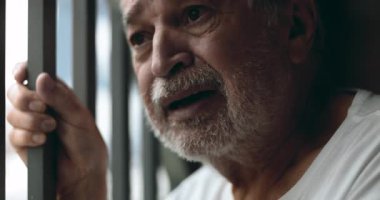 Pencere parmaklıklarını tutan yaşlı bir adam, acıdan gözleri kocaman, evde dayanılmaz kayıplar ve yalnızlıkla yüzleşirken sessizce çığlık atıyor.