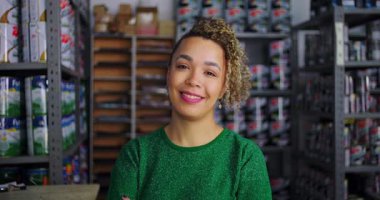 Bir hırdavatçıda gülümseyen kadın girişimci, arka planda boya ve malzeme rafları ile güven ve yakınlık yayıyor.