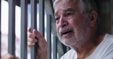 Parmaklıkları çeken yaşlı bir adam, gözü yaşlı gözlerle yukarıya bakıyor, umutsuzluk, duygusal çöküntü, üzüntü, yaşlanma ve zihinsel sağlık sorunları tarafından tüketiliyor.