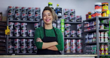 Tezgahın arkasında duran dost canlısı hırdavatçı, kollarını kavuşturmuş, güvenle gülümsüyor, etrafı boya ve malzemelerle dolu renkli raflarla çevrili.