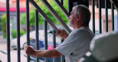 Metal parmaklıkların arkasında duran yaşlı bir adam endişeli bir ifadeyle dışarı bakıyor, iki eliyle parmaklıklara tutunuyor, yaşlılıktan izole edilmiş mahalleyi gözetliyor.