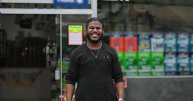 Gülümseyen genç bir siyah adam perakende mağazasının önünde duruyor, güvenini, pozitifliğini ve profesyonel ve müşteri dostu bir çevrede misafirperver tavrını yansıtıyor.