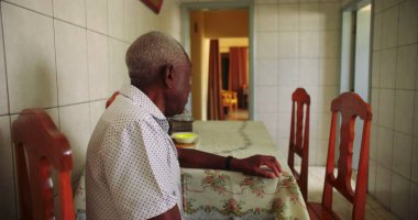 Sessiz bir mutfakta tek başına yemek masasında oturan yaşlı Afrikalı Amerikalı adam, huzur dolu bir ev ortamında yalnızlığı ve içselliği yansıtıyor.